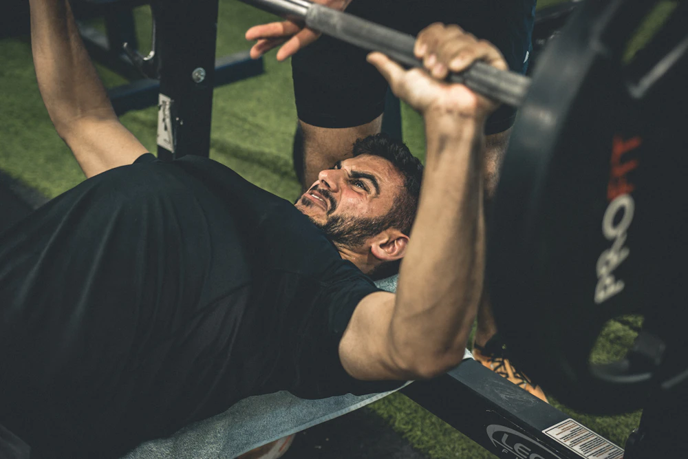 A man straining through a bench press while a spotter stands behind him, hands ready under the bar